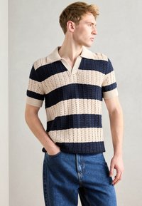 Cămașă polo tricotată cu guler în V, având dungi orizontale bleumarin și crem, mâneci scurte și un model texturat. Asortată cu blugi albaștri.