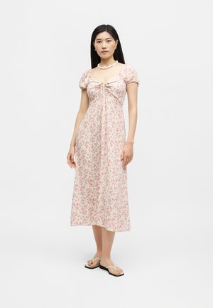 Γυναίκα που στέκεται, φοράει ροζ midi φόρεμα με φλοράλ σχέδιο, με φουσκωτά μανίκια, τετράγωνο ντεκολτέ και μπεζ σανδάλια, σε απλό λευκό φόντο.