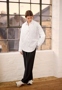Hvid skjorte med knapper og en brystlomme, suppleret med løse sorte bukser og lyse sneakers, sat mod et stort vindue.