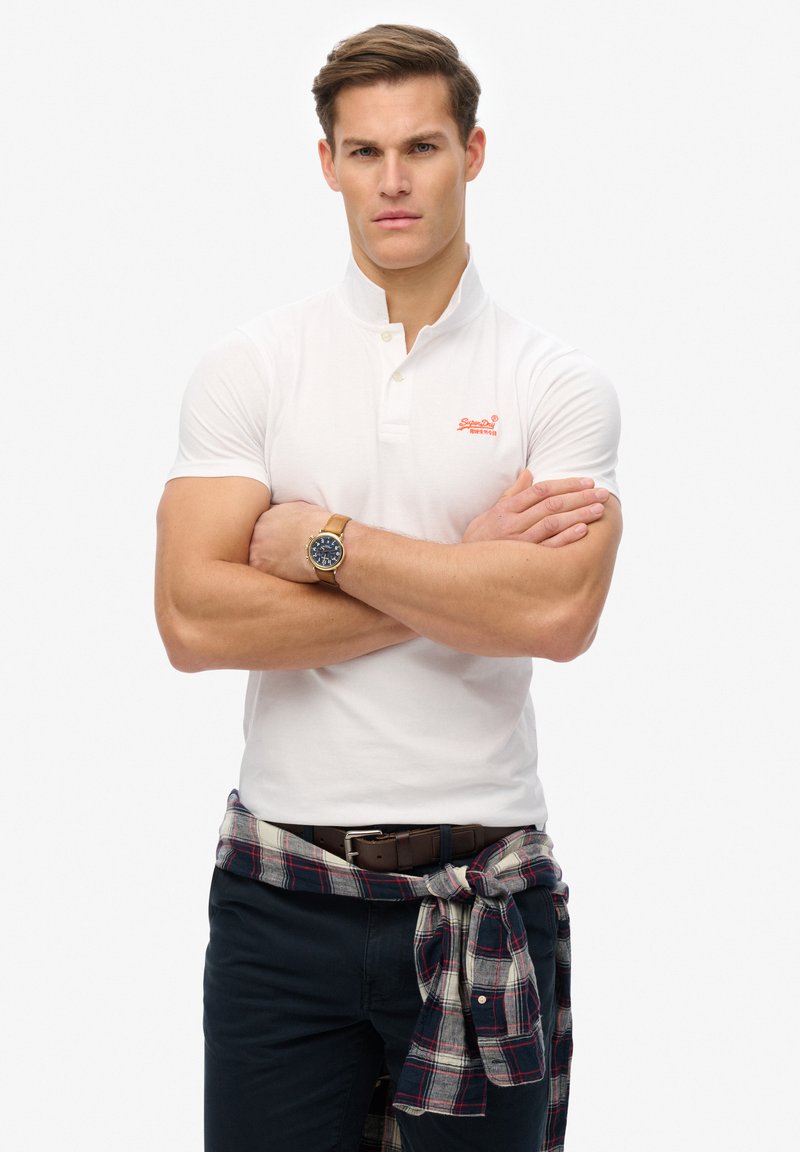 Man met gekruiste armen, gekleed in een witte korte mouwen polo, een geruite shirt om de taille geknoopt, marineblauwe broek en een horloge, tegen een effen witte achtergrond.