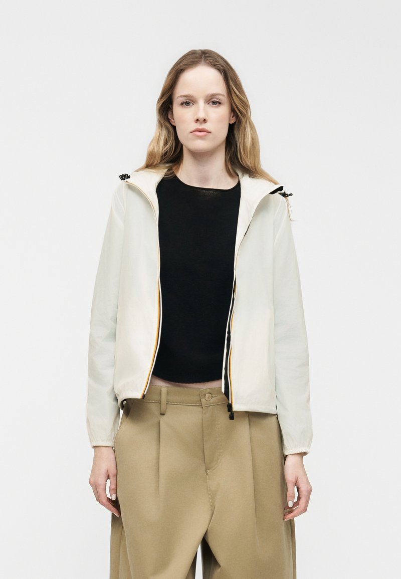 Γυναίκα με μακριά μαλλιά, ντυμένη με λευκό φούτερ με φερμουάρ και κουκούλα, μαύρο τοπ και φαρδιά μπεζ παντελόνια, στέκεται μπροστά σε ένα απλό φόντο.