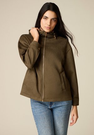 Žena s dlouhými tmavými vlasy, oblečená v olivově zelené bundě na zip a modrých džínách s bílými tečkami, stojící před jednobarevným béžovým pozadím.