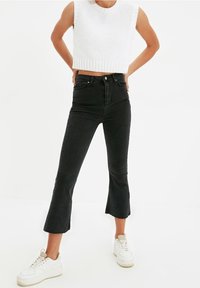 Svarta högmidjade jeans med utsvängd fåll, kombinerade med en vit ärmlös stickad kort topp och vita sneakers. Mjuk textur.