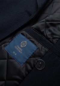 Dunkler Wollmischblazer mit Innenfutter aus schwarzem Steppmaterial. Blaue Etikette mit "Ombre Heritage" in Gold. Klare Knopfdetails sichtbar.