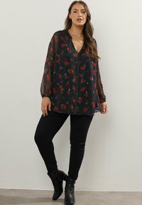 Svart blommig blus med genomskinliga långa ärmar, djup V-ringning och dekorativ nitar längs mitten. Buren med svarta tajta byxor.