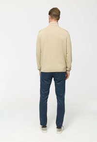 Beige kötött pulcsi magas gallérral, sötétkék nadrággal párosítva. Egyszerű dizájn, puha textúrával. Lábak bézs sportcipőben.