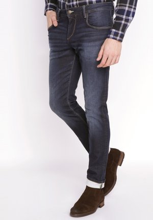 Donkerblauwe spijkerbroek met een slim fit, met contrasterende gele stiksels en een vijf-pocket ontwerp. Opgerolde zoom boven bruine suède laarzen.