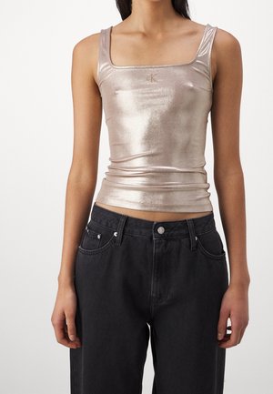 Frau trägt ein metallisch beiges ärmelloses Crop-Top mit quadratischem Ausschnitt und schwarze High-Waist-Jeans vor weißem Hintergrund.