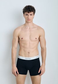 Černé boxerky s bílým logem Calvin Klein na pásku, přiléhavý střih a hladká tkanina, předváděné modelem na světlém pozadí.