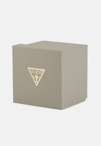 Scatola grigia strutturata con un logo triangolare dorato sul fronte, che presenta il nome del marchio e un motivo a punto interrogativo.