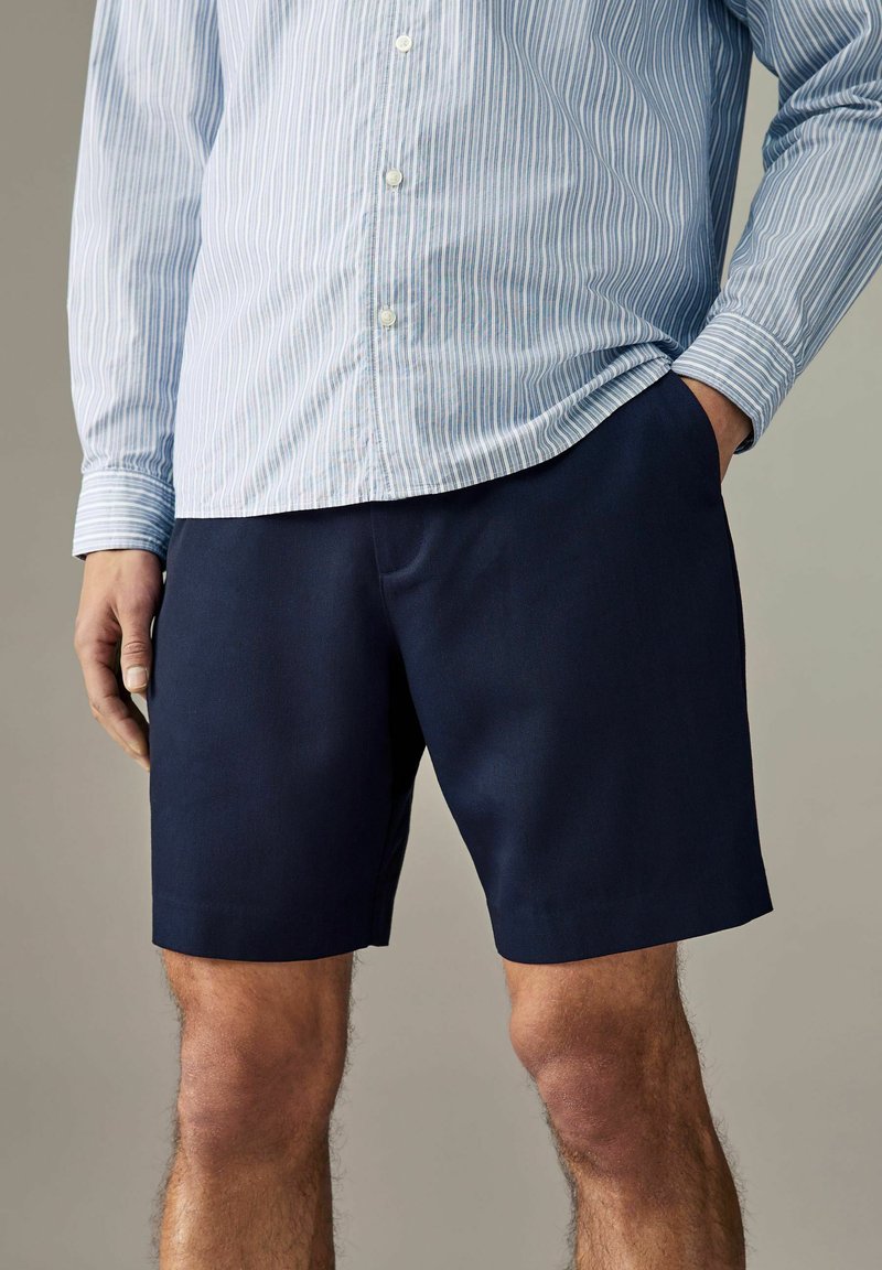 Pantaloni scurți de culoare albastru marin, dintr-un material ușor, cu o croială relaxată și buzunare laterale. Asortați cu o cămașă cu dungi fine, cu nasturi.