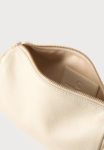 Κρέμ δερμάτινη θήκη με υφή και χρυστό φερμουάρ που είναι μερικώς ανοιχτό, αποκαλύπτοντας λείο υφασμάτινο εσωτερικό και μια μικρή ραμμένη δερμάτινη ετικέτα στο εσωτερικό.