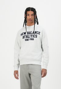 Gaiši pelēks kokvilnas sporta krekls ar tumši zilu tekstu "New Balance Athletics Since 1906." Iekļauj apaļu kakla izgriezumu un ribotas aproces.