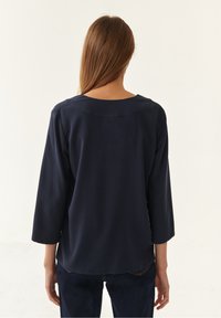 Un haut bleu marine, ample, avec des manches trois-quarts, un col rond et une texture lisse, porté avec un jean foncé.