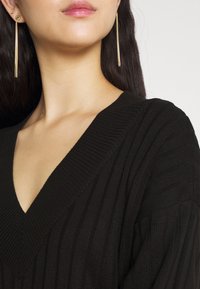 Svart ribbat stickat linne med V-ringning, med texturerade linjer och en avslappnad passform. Guldhängande örhängen synliga, som framhäver ringningen.