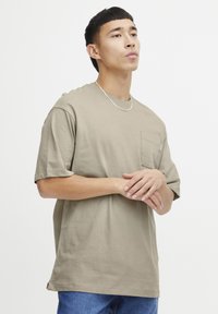 Khaki tričko s krátkým rukávem z bavlny, s náprsní kapsou a uvolněným střihem. Hladký povrch s kulatým výstřihem.