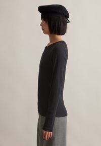 Schwarzes Langarmoberteil mit Rundhalsausschnitt, kombiniert mit einem grauen Streifenshirt. Ein schwarzer Hut wird auf dem Kopf getragen. Seitenansicht.