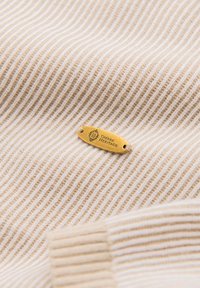 Tissu côtelé beige et blanc avec une petite étiquette en métal doré attachée, gravée de "OMBRE HERITAGE" et d'un logo décoratif.