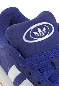 Lilla semsket sneaker med hvite skinnstriper og teksturerte snørebånd. Rund logoakkent på tungen med et bladdesign.