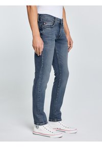 Személy karcsúsított kék farmernadrágot, fehér magas szárú tornacipőt és fehér inget visel, és egyszerű világos háttér előtt áll.