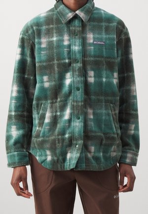 Hombre que lleva una chaqueta de forro polar a cuadros verde y gris con botones de presión y bolsillos laterales, combinado con pantalones marrones.