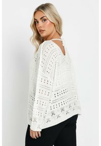 Vit långärmad stickad tröja med ett öppet mönster, andningsbar textur och en knytning bak. Har en avslappnad passform och fåll.