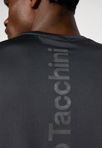 Μαύρο αθλητικό μπλουζάκι με λείο υφή και κάθετο λογότυπο σε γυαλιστερή εκτύπωση κατά μήκος της πλάτης. Κοντά μανίκια και στρογγυλή λαιμόκοψη.