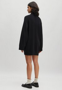 Schwarz oversize Strickkleid mit langen Ärmeln, geripptem Textur und hohem Ausschnitt. Tragen mit schwarzen Schuhen und hellen Socken.