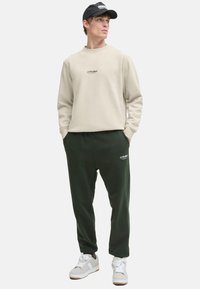 Beige sweatshirt with logo, paired with dark green joggers featuring side pockets. Worn with white and gray sneakers and a black cap.
