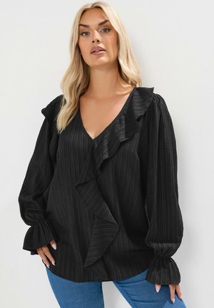 Schwarze Bluse mit Struktur, tiefem V-Ausschnitt, Rüschenakzenten und voluminösen Ärmeln, kombiniert mit blauen Jeans. Weicher, leichtes Material.