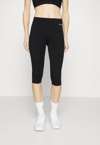 Black capri leggings made of stretchy fabric, featuring a smooth texture and a small logo on the waistband. Worn with white athletic socks and shoes.
