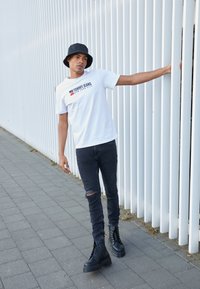 T-shirt bianco Tommy Jeans con logo, jeans skinny neri strappati e stivali piattaforma neri con lacci, posizionati davanti a una parete a strisce verticali bianche.