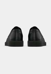 Chaussures en cuir noir avec une texture lisse, une forme arrondie et un petit talon en caoutchouc noir. Elles présentent un design à enfiler et un style minimaliste.
