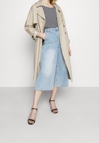 Beige trench coat přes černobíle pruhovaný top, spárovaný s světle modrými culoty a černými sandály na tenkých řemíncích. Hladké textury látek.