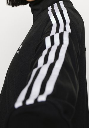 Osoba ubrana w czarną kurtkę sportową Adidas z białymi trzema paskami na rękawie i logo na piersi, wyciągająca ramię do przodu.