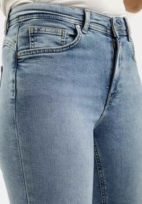 Jasnoniebieskie dżinsy mają dopasowany krój, pięć kieszeni oraz subtelne wyblaknięcia na materiale. Gładka faktura i klasyczne szwy.