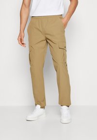 Pantalons cargo kaki avec une taille élastique, deux poches latérales et une coupe fuselée. Associés à des baskets blanches.