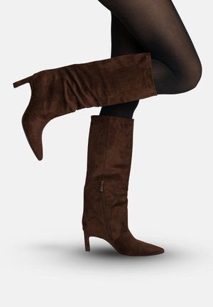 Bruine suède kniehoge laarzen met ritssluiting aan de zijkant en slanke hakken, gedragen met zwarte panty's op gebogen benen.