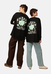 Schwarze Langarm-T-Shirts zeigen einen verspielten Grafikdruck mit grünen Akzenten auf dem Rücken. Models tragen braune und hellblaue Hosen und stehen sich gegenüber.