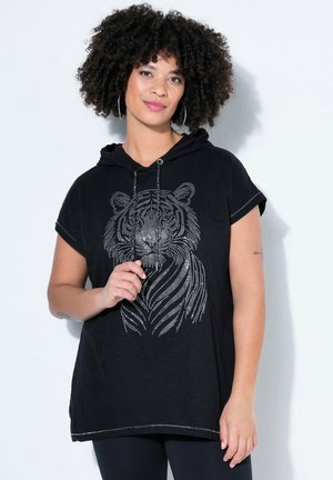 Woman with curly hair wearing hoop earrings and a black short-sleeve hoodie with a dotted tiger face design, standing against a plain white background.