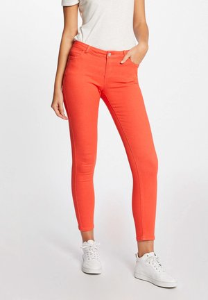 Femme portant un pantalon ajusté orange vif avec des poches, des baskets blanches et un haut blanc, debout devant un fond clair uni.