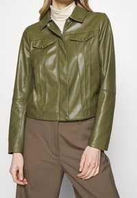 Veste en simili cuir vert olive avec un col, fermeture éclair et poches poitrine. Présente des coutures verticales et des manches ajustées.