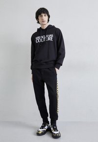 Černá mikina s kapucí a bílým logem "VERSACE JEANS COUTURE", spárovaná s černými kalhotami s žlutým bočním pruhem. Boty mají výraznou grafiku.