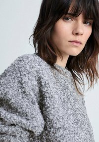 Maglione grigio, con texture e una vestibilità ampia, dal finish morbido e peloso. Il modello ha i capelli lunghi fino alle spalle e un'espressione neutra.