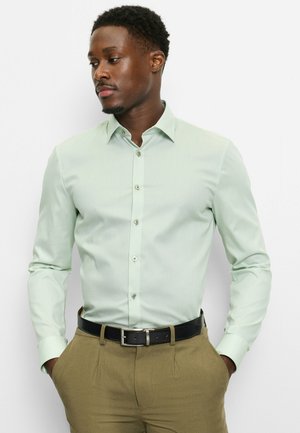 Mann in hellgrünem Hemd mit Knöpfen und beigen Hosen mit schwarzem Gürtel, Hände in den Taschen, blickt nach links vor weißem Hintergrund.