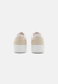 Cream sneakers with embossed logo on the back, patterned design on the heel, and white rubber sole. Features yellow accents on sides.