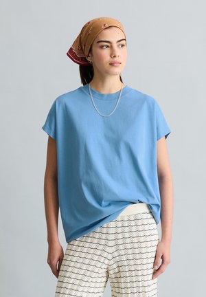 Młoda kobieta w jasnoniebieskim topie bez rękawów, kremowych spodniach w kratę, brązowej wzorzystej chustce na głowie i srebrnym naszyjniku z łańcuszkiem, zwrócona bokiem.