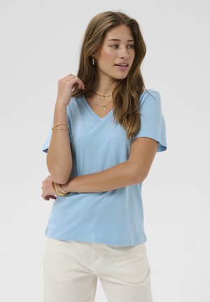 Kvinde med langt brunt hår iført lysblå V-udskåret T-shirt og hvide bukser, accessoriseret med guldbøjler, halskæder og øreringe.