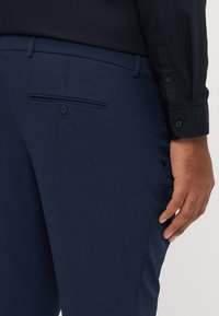 Marineblaue Anzughose mit schmaler Passform, glatter Textur und einer Gesäßtasche mit Knopf. Kombiniert mit einem schwarzen Langarmshirt.