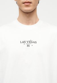 Άτομο που φοράει λευκό μπλουζάκι με την επιγραφή «LAS VEGAS REPLAY 81» σε μαύρο, τοποθετημένη στο κέντρο του στήθους.